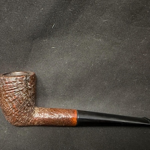 May include: A brown tobacco pipe with a textured bowl and a dark, curved stem. The pipe has a classic design, with a wooden bowl and a black mouthpiece. The pipe is approximately 6 inches long.
