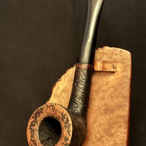 Pode incluir: Um cachimbo de tabaco com fornilho de madeira de urze escura e texturizada e piteira de acrílico preto liso. O cachimbo está posicionado na vertical contra um suporte de madeira polida castanho-claro com veios e bordos irregulares.