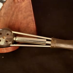 May include: A smoking pipe featuring a dark brown, rounded bowl with four small circular holes on the front face. The stem is constructed from a central metallic cylinder flanked by two thin metal rods, leading to a dark brown, cylindrical mouthpiece.