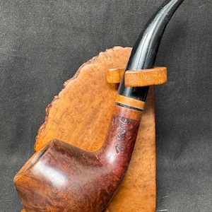 May include: A curved, brown smoking pipe with a black stem, resting on a wooden stand. The pipe has a rounded bowl and a curved stem. The wooden stand is light brown and has a natural wood grain pattern. The pipe has the text "Mr. Brog" engraved on the side.