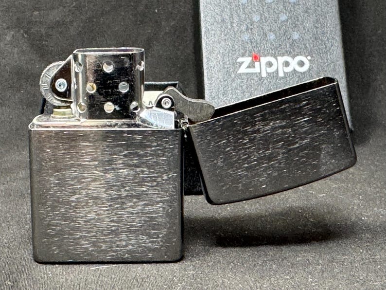 May include: A brushed metal Zippo lighter with the lid open, showing the inside. The lighter is rectangular with rounded edges and a textured surface. The Zippo logo is visible on the packaging in the background.