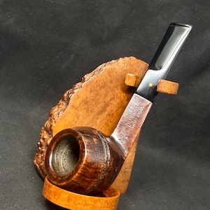 May include: A wooden pipe with a dark, curved stem and a round bowl. The bowl is a darker brown with a textured surface. The stem is black with a gold dot. The pipe rests on a wooden stand.
