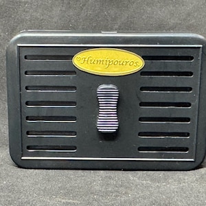 May include: A black rectangular box with horizontal slots and a gold oval label that reads "Humipouros." The box has a silver and blue striped handle. The item appears to be a storage container.