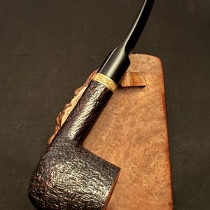 Puede incluir: Una pipa de tabaco con un hornillo oscuro, texturizado y rústico, y una boquilla negra lisa. Una banda de metal dorado con un patrón grabado conecta el hornillo con la boquilla. La pipa descansa sobre un soporte de madera de color claro.