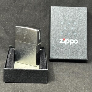 May include: A silver Zippo lighter sits in a black box with a velvet lining. The lighter has a rectangular shape with rounded edges and a brushed metal finish. The Zippo logo is printed on the box.