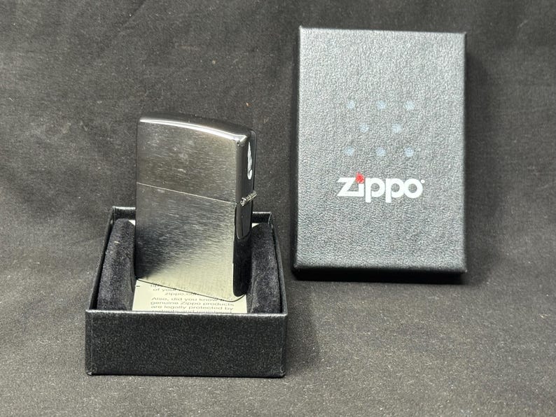 May include: A silver Zippo lighter sits in a black box with a velvet lining. The lighter has a rectangular shape with rounded edges and a brushed metal finish. The Zippo logo is printed on the box.