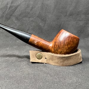 May include: A brown and black tobacco pipe with a curved design, resting on a small, tan leather stand. The pipe has a dark, glossy mouthpiece and a rounded bowl. The stand has a brass-colored button.