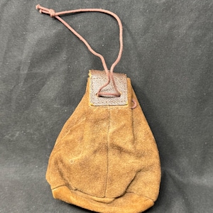 Puede incluir: Una bolsa de gamuza marrón con cierre de cordón. La bolsa tiene un fondo redondeado y una pequeña solapa rectangular de cuero en la parte superior donde se adjunta el cordón. El cordón es de color marrón claro.