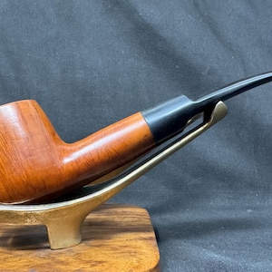 Könnte beinhalten: Eine Tabakpfeife aus Holz mit einem gebogenen schwarzen Mundstück und einem braunen Kopf. Sie steht auf einem goldfarbenen Ständer, der auf einem Holzsockel platziert ist. Der Hintergrund ist dunkel.