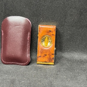 May include: A vintage lighter with a brown wood grain finish and gold accents, next to a burgundy leather case. The lighter features a gold oval design in the center. The case is rectangular with rounded edges.