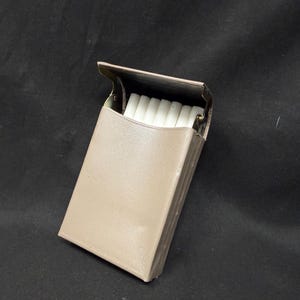May include: A tan cigarette case, open to reveal a stack of white cigarettes. The case is rectangular with a hinged lid and a textured surface. The background is a dark, solid color.