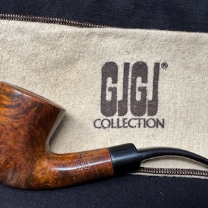 May include: A brown and black pipe with a curved stem, resting on a beige cloth with the text "GJGJ COLLECTION". The pipe has a polished finish and a dark, curved mouthpiece. The wood grain is visible.