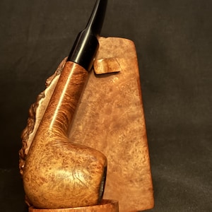 May include: A tobacco pipe with a polished light brown wood grain bowl and a smooth black stem, resting on a matching wooden stand that features a textured, natural burl wood finish on its side and a flat base for stability.