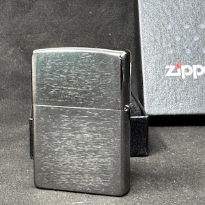 May include: A silver Zippo lighter with a brushed metal finish. The rectangular lighter has a textured surface. The Zippo logo is displayed on the box in the background.