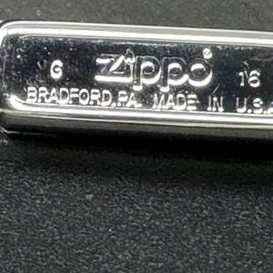 May include: A chrome Zippo lighter with the text "Zippo BRADFORD, PA. MADE IN U.S.A." The rectangular lighter has rounded edges and a shiny finish. The lighter is on a black surface.