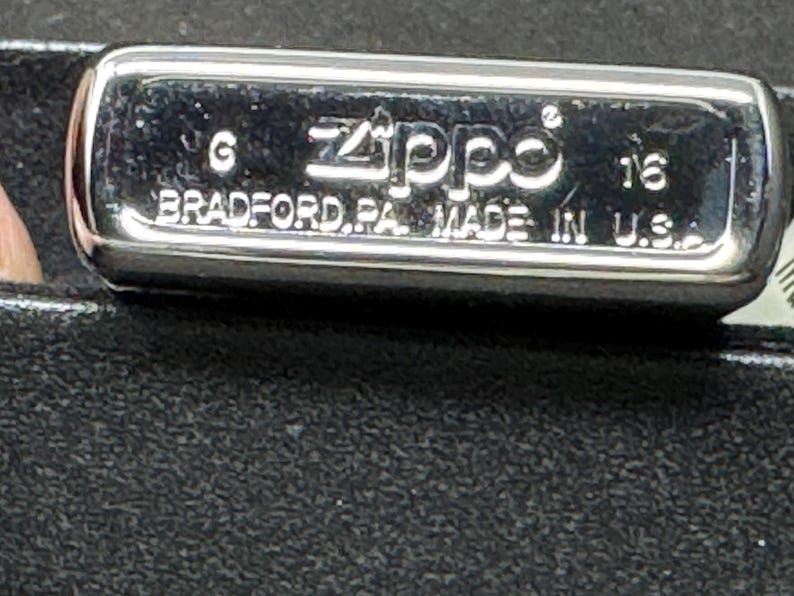 May include: A chrome Zippo lighter with the text "Zippo BRADFORD, PA. MADE IN U.S.A." The rectangular lighter has rounded edges and a shiny finish. The lighter is on a black surface.