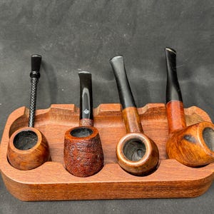 May include: Four wooden tobacco pipes of varying shapes and sizes rest in a wooden pipe stand. The pipes have dark stems and bowls, with a range of wood grains and finishes. The stand is a warm brown color.