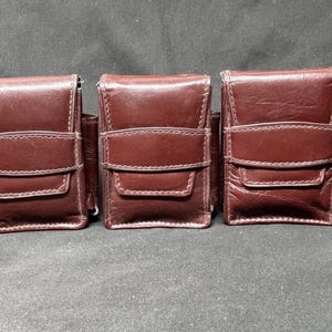 May include: Three brown leather cases with a rectangular shape and a flap closure. Each case has a stitched design and a small pocket on the side. The cases are arranged side by side on a dark surface.