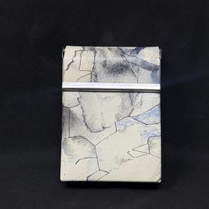 May include: A rectangular case with a silver metal clasp. The case has a white background with a blue and gray abstract design. The case is closed and appears to be made of metal.