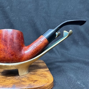 Peut inclure: Une pipe à tabac avec un foyer brun rougeâtre et un tuyau noir, posée sur un support doré. La pipe porte l'inscription "LAS VEGAS". Le support est posé sur une base en bois.