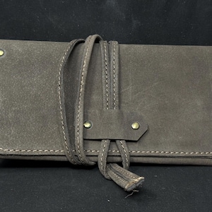 May include: A gray leather roll-up case with a strap closure. The case is rectangular with rounded corners and brass rivets. White stitching accents the edges and strap. The strap is wrapped around the case and tied with a tassel.