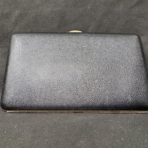Pode incluir: Uma clutch retangular preta com uma superfície texturizada. A clutch tem uma estrutura de metal e um fecho de encaixe. O design é simples e elegante, adequado para várias ocasiões.