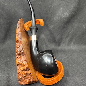 May include: A black smoking pipe with a curved stem, resting on a wooden stand. The pipe has a silver band and a glossy finish. The stand is made of carved wood with a natural finish, providing support for the pipe.