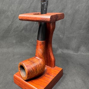 May include: A wooden tobacco pipe with a curved bowl and a black stem, resting on a matching wooden stand. The stand has a square base and a curved support, with a slot to hold the pipe's stem. The wood has a reddish-brown finish.