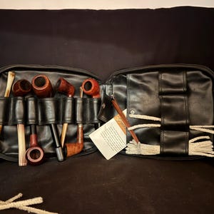 May include: A black leather tobacco pipe carrying case featuring multiple slots for wooden pipes and a dedicated section for pipe cleaners. The case is open, displaying several brown wooden pipes of varying shapes and sizes, along with white pipe cleaners.
