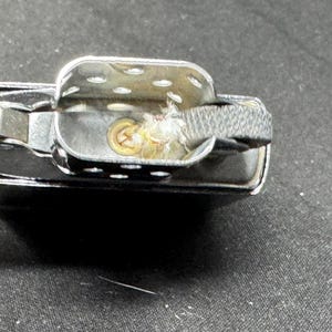 May include: Close-up of a chrome lighter, open to show internal parts. The design includes a hinged lid, a fuel chamber, and a wick. The wick is surrounded by a cotton-like material.