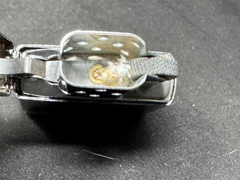 May include: Close-up of a chrome lighter, open to show internal parts. The design includes a hinged lid, a fuel chamber, and a wick. The wick is surrounded by a cotton-like material.