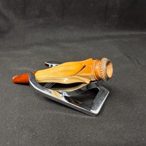 May include: A wooden pipe with a reddish-brown stem and a light-colored body, resting on a chrome-colored metal stand. The pipe's bowl has a textured, dark brown rim. The stand has a triangular shape.