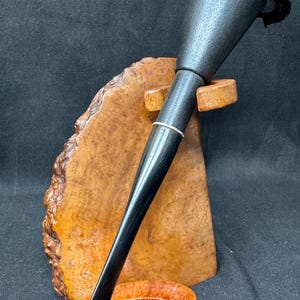 May include: A black pipe with a silver band, resting on a wooden stand. The pipe has a conical bowl and a long, slender stem. The stand is made of a light-colored wood with a natural grain pattern. The background is dark.