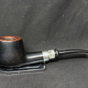 Puede incluir: Una pipa negra con boquilla curva y banda plateada. El cuenco es marrón oscuro con interior marrón claro. La pipa descansa sobre un soporte plateado sobre un fondo oscuro.