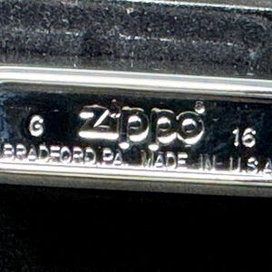 May include: A close-up of a chrome Zippo lighter. The lighter features the Zippo logo and the text "BRADFORD PA. MADE IN U.S.A." embossed on the surface. The number "16" is also visible.