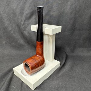 May include: A classic tobacco pipe with a dark brown stem and a reddish-brown bowl, resting on a white stand. The pipe's bowl has a visible grain pattern. The stand is rectangular with a curved indentation to hold the pipe.