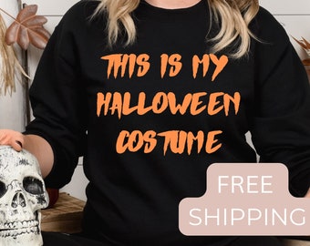 May include: Black sweatshirt with the text "This is my Halloween costume" in orange lettering. The sweatshirt is being worn by a person with long blonde hair. The text "FREE SHIPPING" is on a pink rectangle.