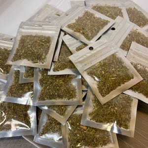 Könnte beinhalten: Eine Ansammlung kleiner, klarer Plastiktüten, gefüllt mit getrockneten, grünen Kräutern. Die Beutel sind versiegelt und in einem Haufen angeordnet, wobei sich einige überlappen. Die Kräuter scheinen fein gehackt zu sein und haben eine natürliche, erdige Farbe.