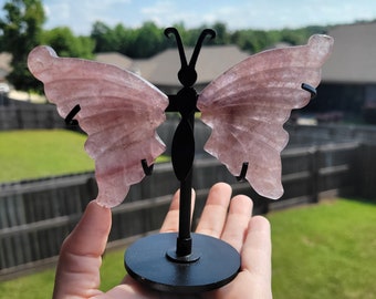 May include: A pink butterfly-shaped crystal on a black metal stand. The crystal is translucent and has a delicate, layered appearance.