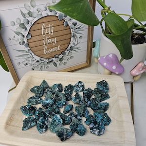 May include: A wooden tray holds several teal-colored chrysocolla mineral specimens. A framed sign with the text "let's stay home" is in the background, along with a potted plant and decorative mushroom figurines.