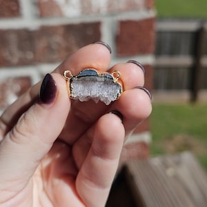 May include: A gold-toned pendant featuring a clear, white, and grey crystal with a rough, textured surface. The crystal is shaped like a cloud and has a gold-toned metal frame.