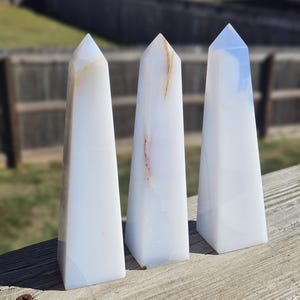 May include: Three white obelisks with subtle brown and blue veining, standing upright on a wooden surface. Each obelisk has a pointed top and a rectangular base. The background is blurred, showing a fence and greenery.