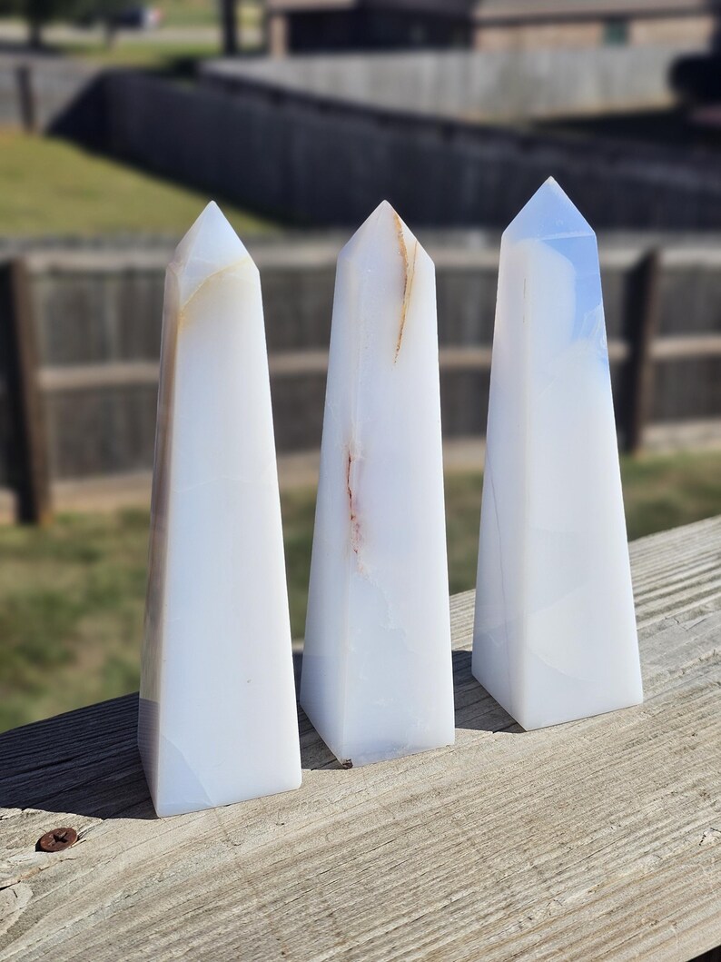 May include: Three white obelisks with subtle brown and blue veining, standing upright on a wooden surface. Each obelisk has a pointed top and a rectangular base. The background is blurred, showing a fence and greenery.