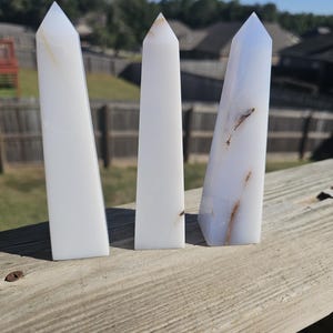 May include: Three white obelisk crystals with pointed tops. The crystals have subtle variations in color and veining, with some showing light brown and blue hues. They stand upright on a wooden surface against a backdrop of trees and a clear blue sky.