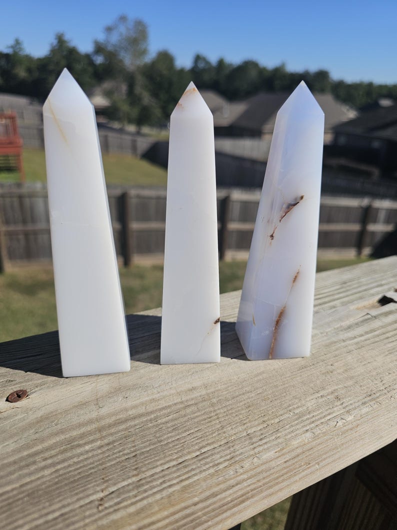 May include: Three white obelisk crystals with pointed tops. The crystals have subtle variations in color and veining, with some showing light brown and blue hues. They stand upright on a wooden surface against a backdrop of trees and a clear blue sky.