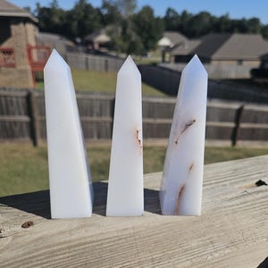 May include: Three white quartz obelisks with pointed tops. The obelisks have subtle brown and orange veining. They are displayed on a weathered wooden surface against a backdrop of a residential neighborhood and a clear blue sky.
