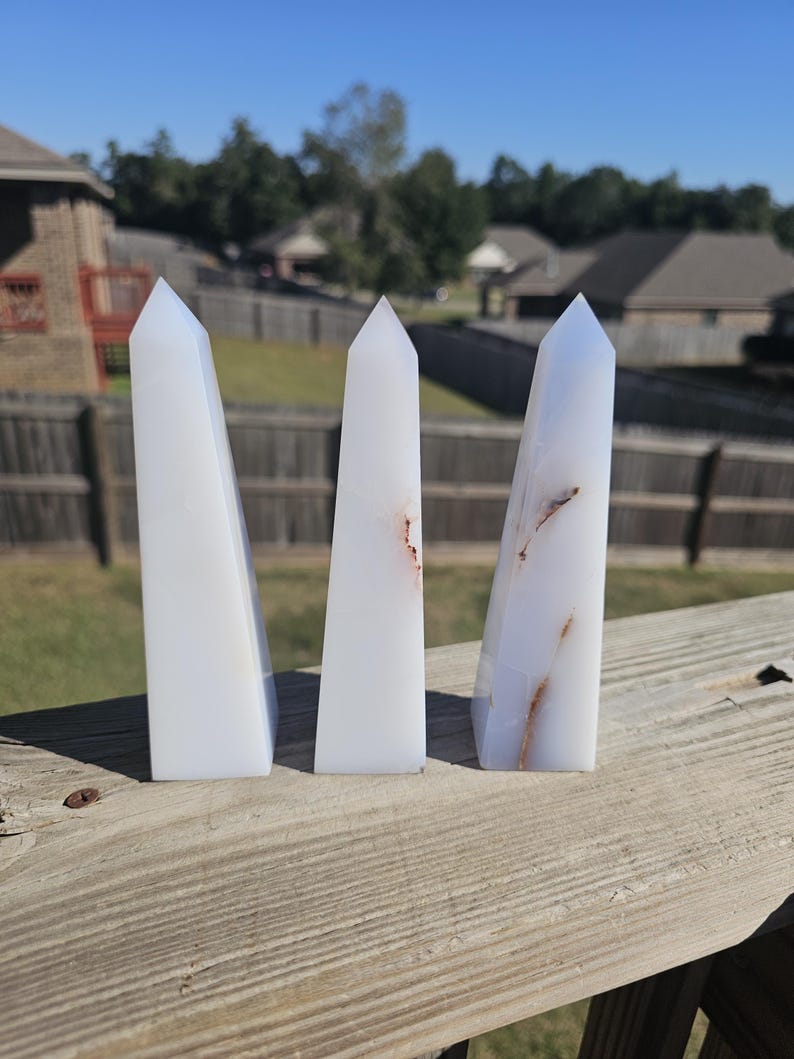 May include: Three white quartz obelisks with pointed tops. The obelisks have subtle brown and orange veining. They are displayed on a weathered wooden surface against a backdrop of a residential neighborhood and a clear blue sky.