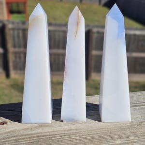 May include: Three white obelisk-shaped crystals with subtle color variations. The crystals are displayed on a weathered wooden surface, with a blurred outdoor background. Each crystal has a pointed top and a rectangular base, showcasing natural stone formations.