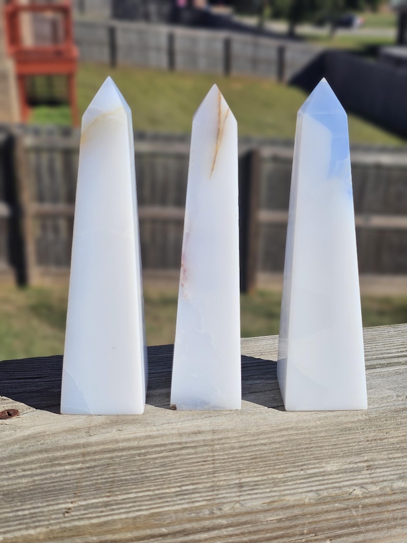 May include: Three white obelisk-shaped crystals with subtle color variations. The crystals are displayed on a weathered wooden surface, with a blurred outdoor background. Each crystal has a pointed top and a rectangular base, showcasing natural stone formations.