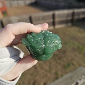 Puede incluir: Una detallada escultura de jade verde de una criatura mítica, posiblemente una rana o un sapo, con intrincadas tallas. La escultura se sostiene en una mano, mostrando su superficie lisa y pulida y sus detalles.